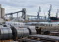 La imagen creada muestra un puerto industrial con acero, materiales a granel y un buque de carga. Es adecuada como imagen de portada para el artículo; es una ilustración y no representa ningún lugar concreto.