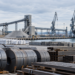 La imagen creada muestra un puerto industrial con acero, materiales a granel y un buque de carga. Es adecuada como imagen de portada para el artículo; es una ilustración y no representa ningún lugar concreto.